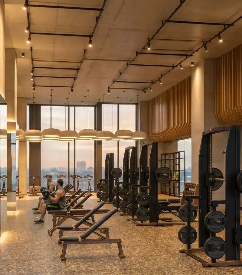 The Uptown gym at sunset, free weights and benches under pendant lights, the city through full-height windows